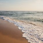 Wellen mit Schaum treffen auf einen ruhigen Sandstrand bei leichtem Sonnenlicht.