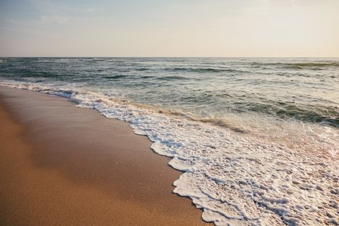 Wellen mit Schaum treffen auf einen ruhigen Sandstrand bei leichtem Sonnenlicht.